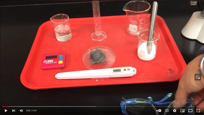 A screenshot of the video students will be watching at the Research It! station. The video is paused on a shot of multiple beakers, tools, and materials on a red tray.